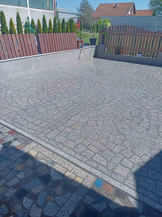 Prostrana dvorišna terasa popločana sitnim komadima sivog granita u lučnim šarama, uz drvenu ogradu.